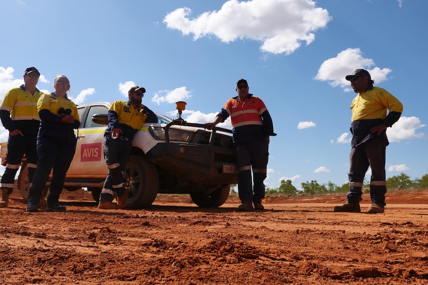 An indigenous person on red earth stands next to a ute surrounded by people in high visibility